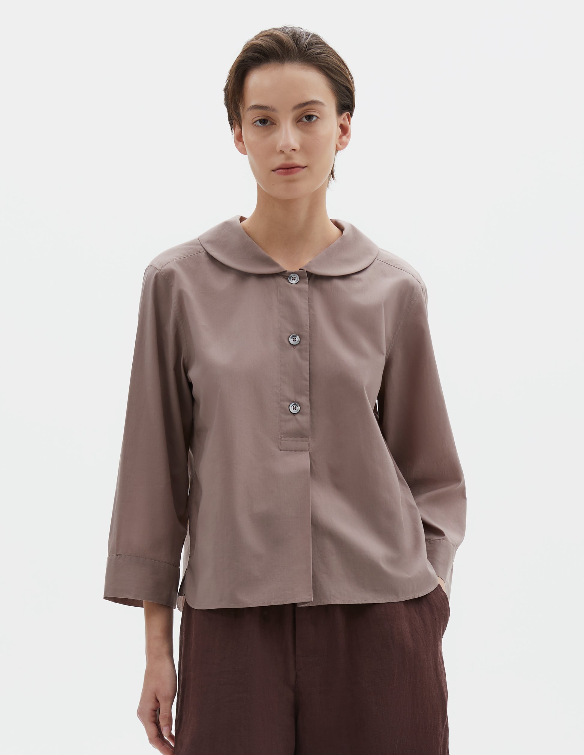 Cropped body image, front view of model wearing pinkish brown pull-on shirt with rounded collar, stitch-through half placket and three quarter sleeves with a turned back cuff detail. Styled with brown shorts.