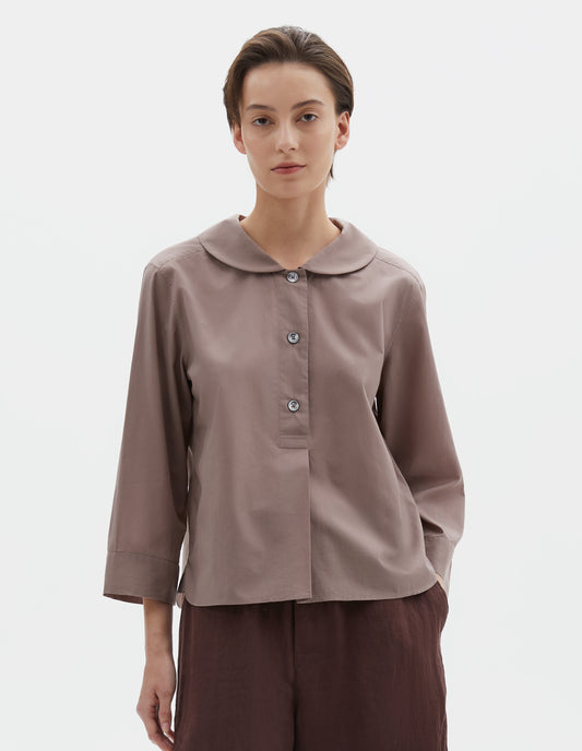 Cropped body image, front view of model wearing pinkish brown pull-on shirt with rounded collar, stitch-through half placket and three quarter sleeves with a turned back cuff detail. Styled with brown shorts.