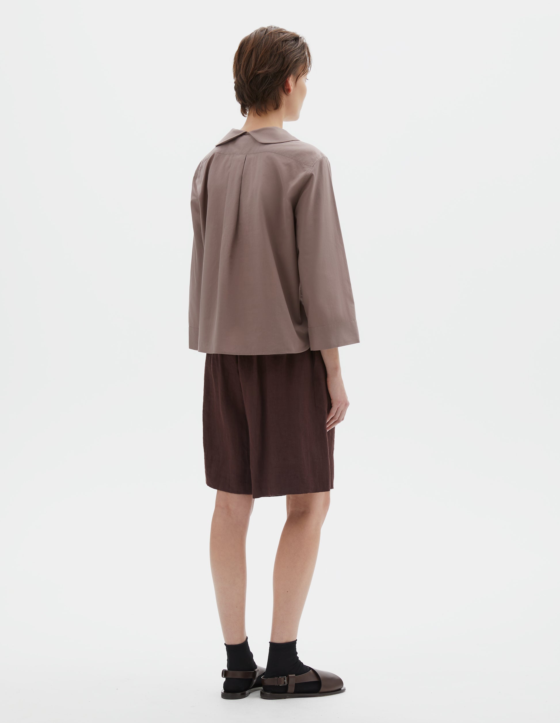 Full body image, back view of model wearing pinkish brown pull-on shirt  with round petal collar detail, box pleats on back yoke, three quarter sleeves with turned back cuff detail. Styled with brown shorts and black socks with dark brown leather sandals.