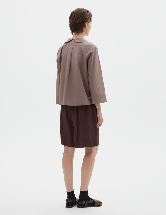 Full body image, back view of model wearing pinkish brown pull-on shirt  with round petal collar detail, box pleats on back yoke, three quarter sleeves with turned back cuff detail. Styled with brown shorts and black socks with dark brown leather sandals.