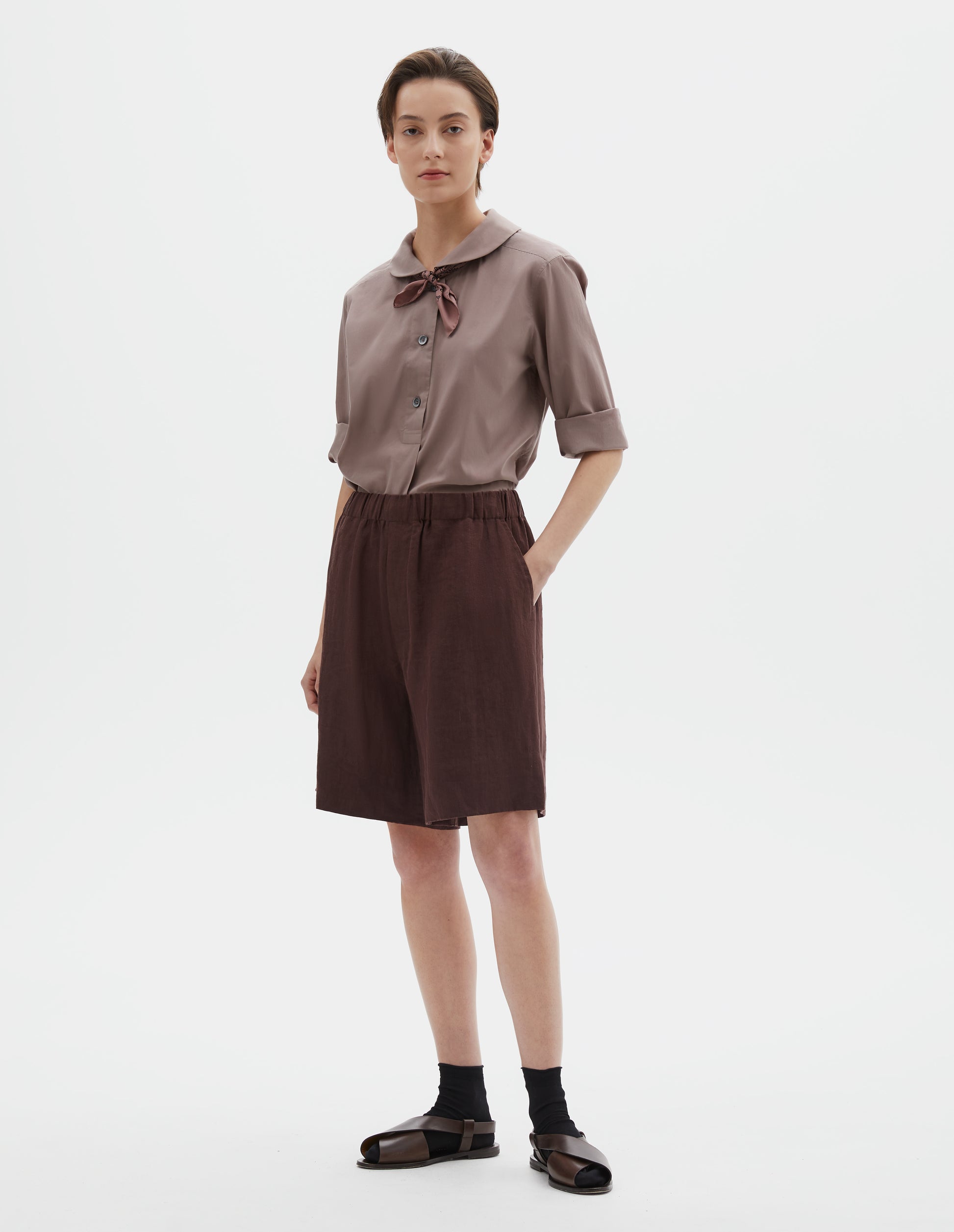Full body image, front view of model wearing reddish brown linen shorts, with elasticated waistband, side entry pockets and deep hem. Styled with pink petal collar shirt tucked in, pink bandana tied under the collar of the shirt, black socks and brown leather sandals.