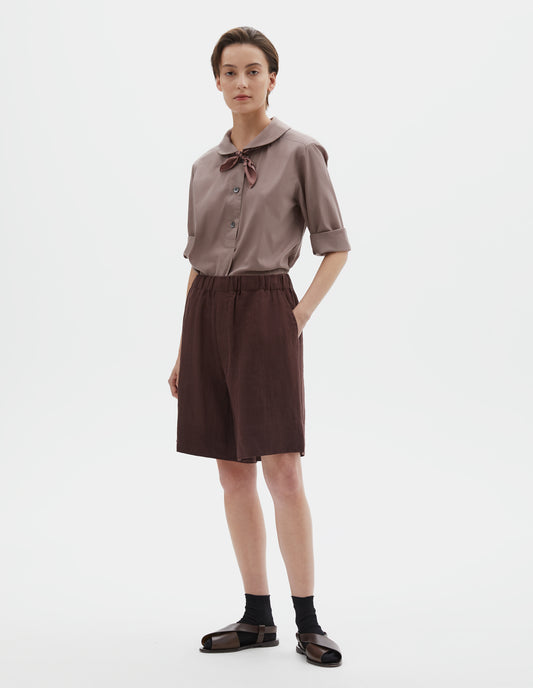Full body image, front view of model wearing reddish brown linen shorts, with elasticated waistband, side entry pockets and deep hem. Styled with pink petal collar shirt tucked in, pink bandana tied under the collar of the shirt, black socks and brown leather sandals.
