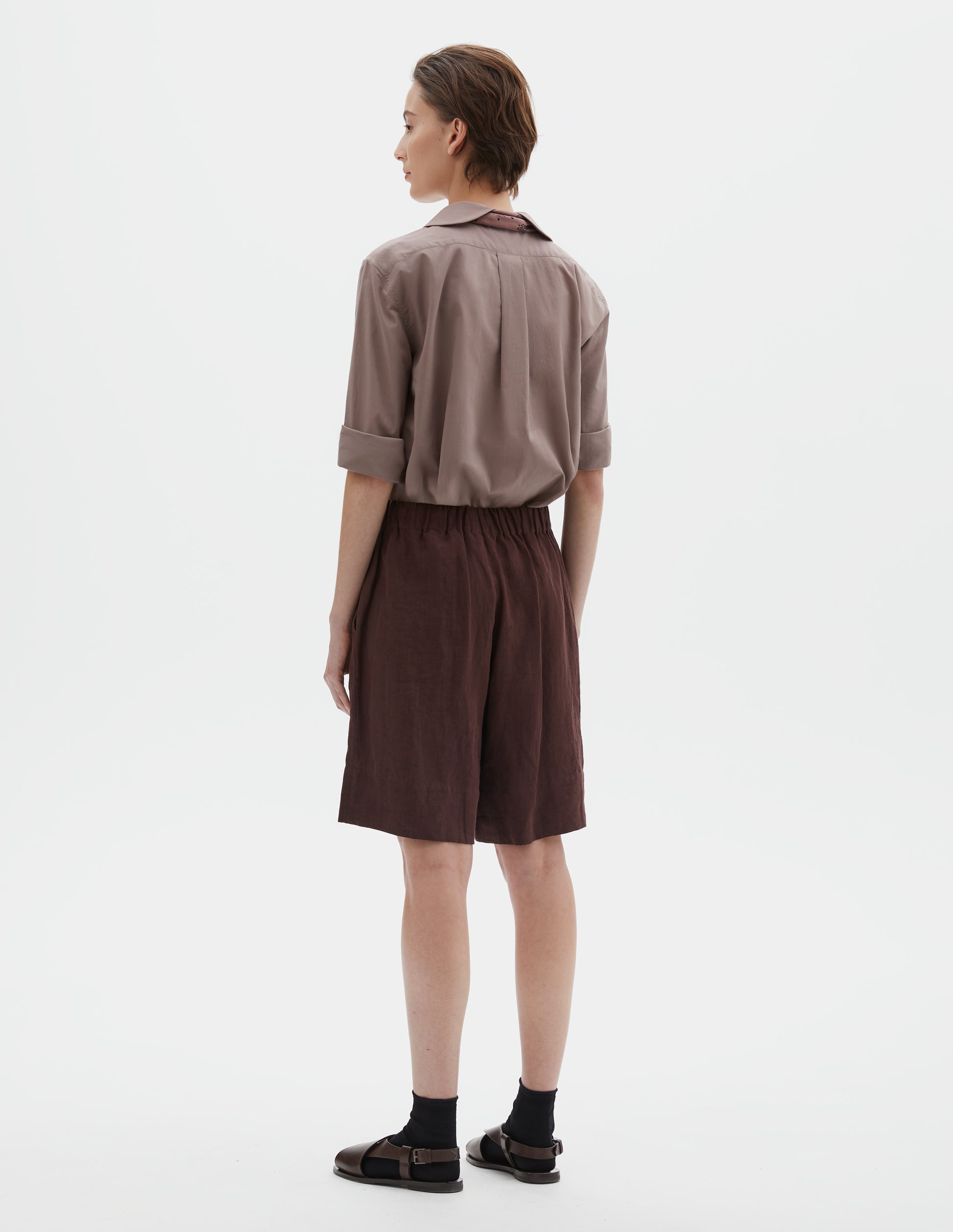 Full body image, back view of model wearing reddish brown linen shorts, with elasticated waistband, side entry pockets and deep hem. Styled with pink petal collar shirt tucked in, pink bandana tied under the collar of the shirt, black socks and brown leather sandals.