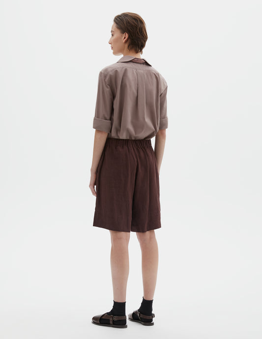 Full body image, back view of model wearing reddish brown linen shorts, with elasticated waistband, side entry pockets and deep hem. Styled with pink petal collar shirt tucked in, pink bandana tied under the collar of the shirt, black socks and brown leather sandals.