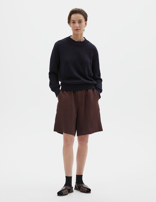 Full body image, front view of model wearing dark navy blue linen and cotton crew neck jumper, with deep rib crew neck, rib cuffs and hem. Styled over pink curve collared shirt, with brownish red shorts, black socks and brown leather sandals.