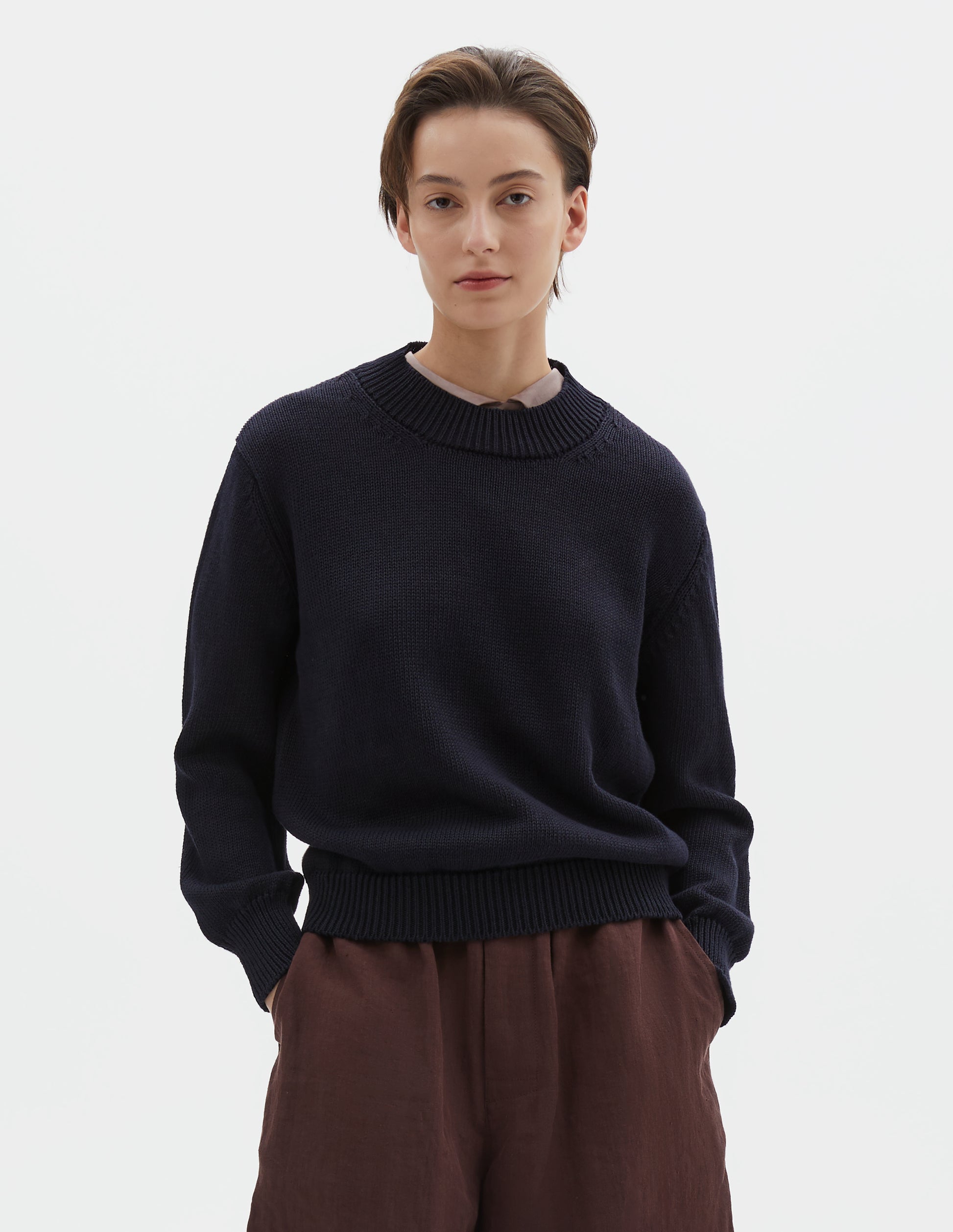 Cropped body image, front view of model wearing dark navy blue linen and cotton crew neck jumper, with deep rib crew neck, rib cuffs and hem. Styled over pink curve collared shirt, with brownish red shorts.