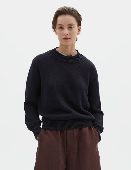 Cropped body image, front view of model wearing dark navy blue linen and cotton crew neck jumper, with deep rib crew neck, rib cuffs and hem. Styled over pink curve collared shirt, with brownish red shorts.