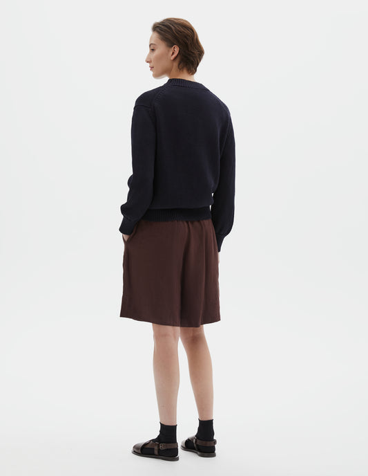 Full body image, back view of model wearing dark navy blue linen and cotton crew neck jumper, with deep rib crew neck, rib cuffs and hem. Styled with brownish red shorts, black socks and brown leather sandals.