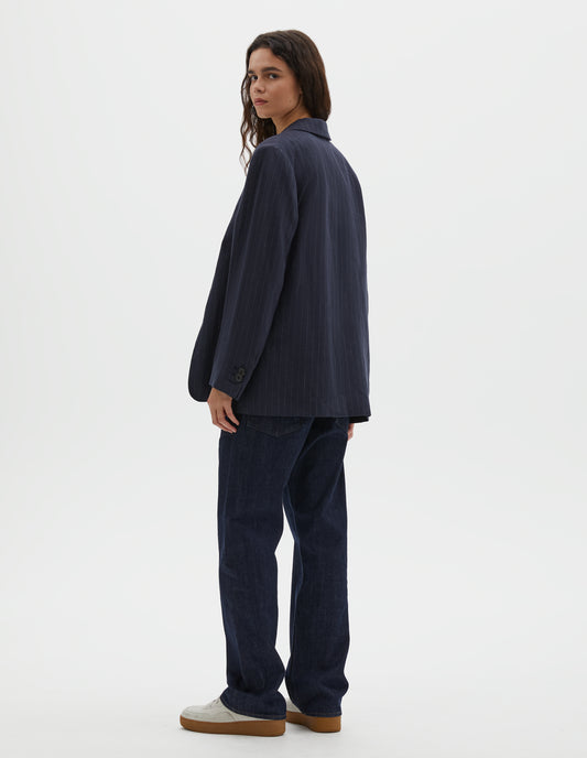 Full body image, back view of model wearing dark navy blue and grey pinstripe linen blazer, with horn button cuffs and back vent. Styled with indigo blue denim jeans and off white suede trainers.