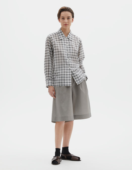 Full body image, front view of model wearing white with black check shirt with a curved collar, side split hem. Styled with black and beige fine stripe shorts and black socks with dark brown leather sandals.