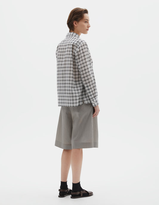 Full body image, back view of model wearing white with black check shirt with pleats on the back yoke, and a side split hem. Styled with black and beige fine stripe shorts and black socks with dark brown leather sandals.