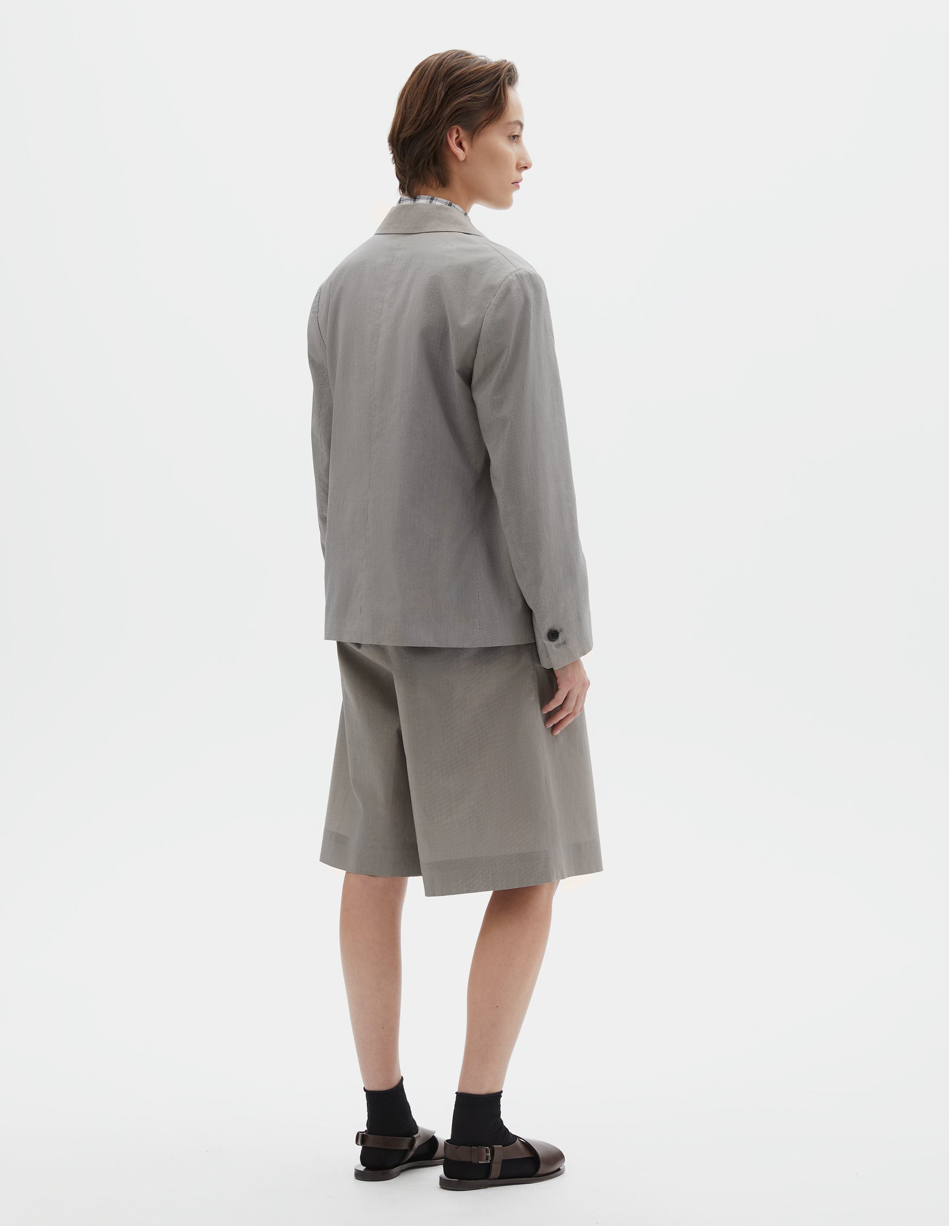 Full body image, back view of model wearing black and cream stripe cotton silk blazer with horn button cuffs. SStyled over black and white checked shirt, with matching black and cream stipe cotton silk shorts, black socks and brown leather sandals.