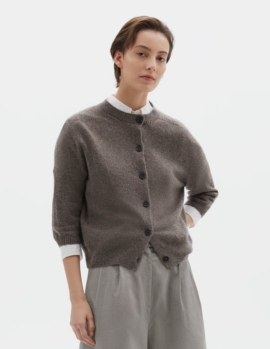 Cropped body image, front view of model wearing greyish beige cashmere cotton cardigan, with elbow length saddle sleeve and large horn buttons. Styled over white cotton shirt, and grey pleated shorts. 