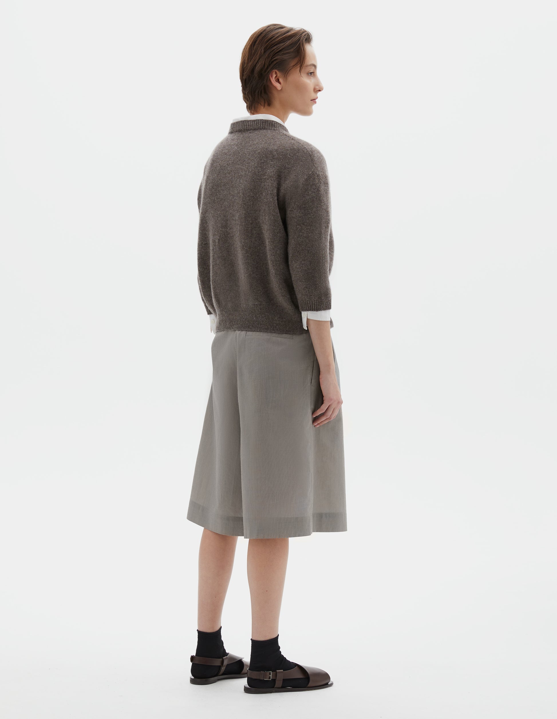 Full body image, back view of model wearing greyish beige cashmere cotton cardigan, with elbow length saddle sleeve. Styled over white cotton shirt, grey pleated shorts, black socks and brown leather sandals.