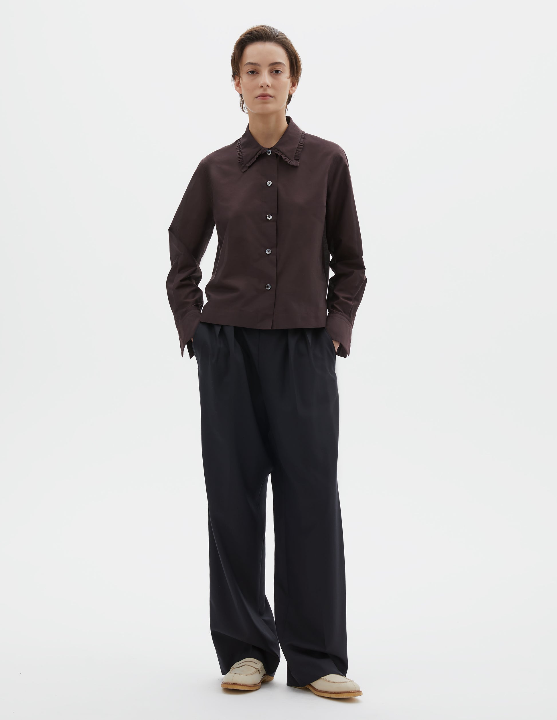 Full body image, front view of model wearing dark red cotton silk frilled collar long sleeve shirt, with mother of pearl button closure. Styled with dark navy wide leg trousers and beige loafers.