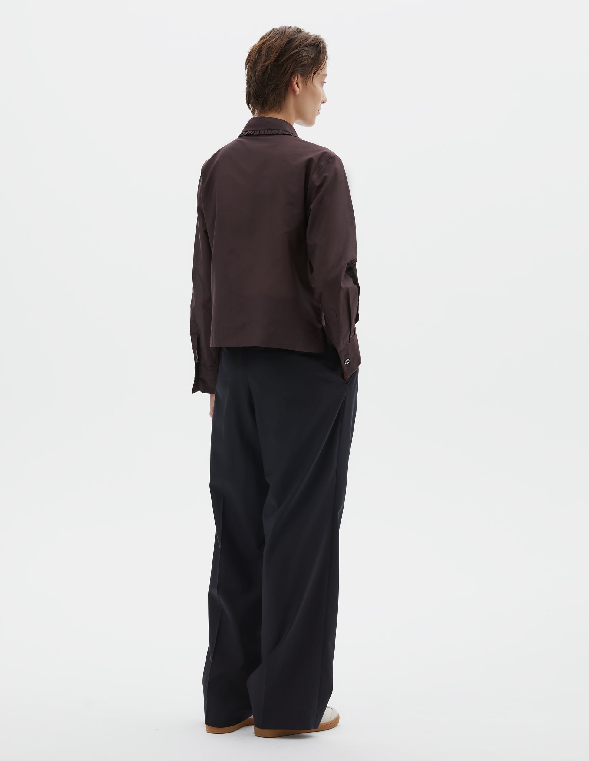Full body image, back view of model wearing dark red cotton silk frilled collar long sleeve shirt, with mother of pearl button cuffs, Styled with dark navy wide leg trousers and beige loafers.