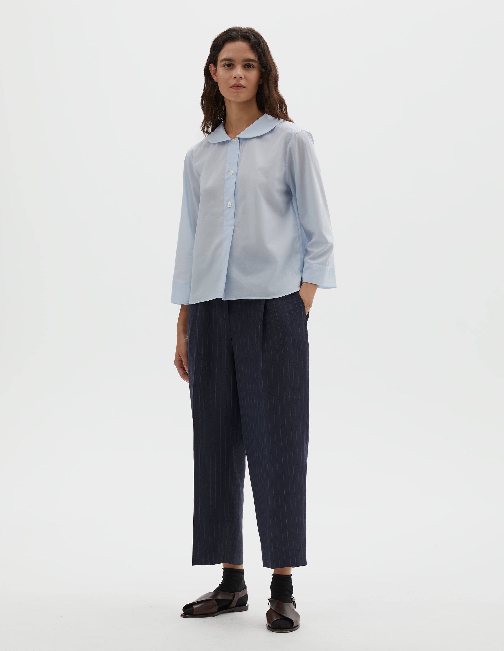 Full body image, front view of model wearing light blue cotton shirt, with round petal collar, stitch through half placket, mother of pearl buttons and three quarter sleeves with turned back cuff detail. Styled with navy blue and grey pinstripe trousers, black ankle socks and brown leather sandals.