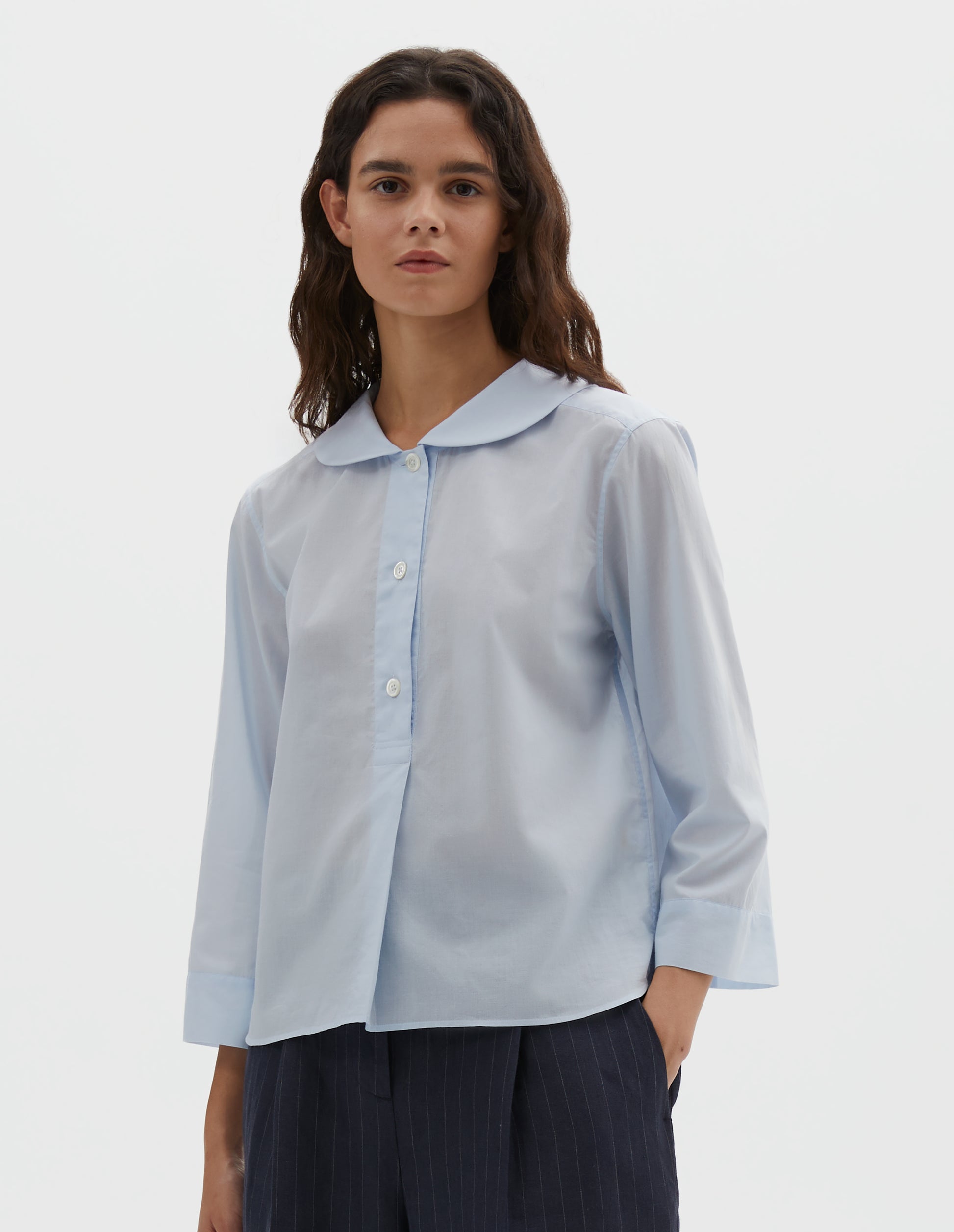 Cropped body image, front view of model wearing light blue cotton shirt, with round petal collar, stitch through half placket, mother of pearl buttons and three quarter sleeves with turned back cuff detail. Styled with navy blue and grey pinstripe trousers.