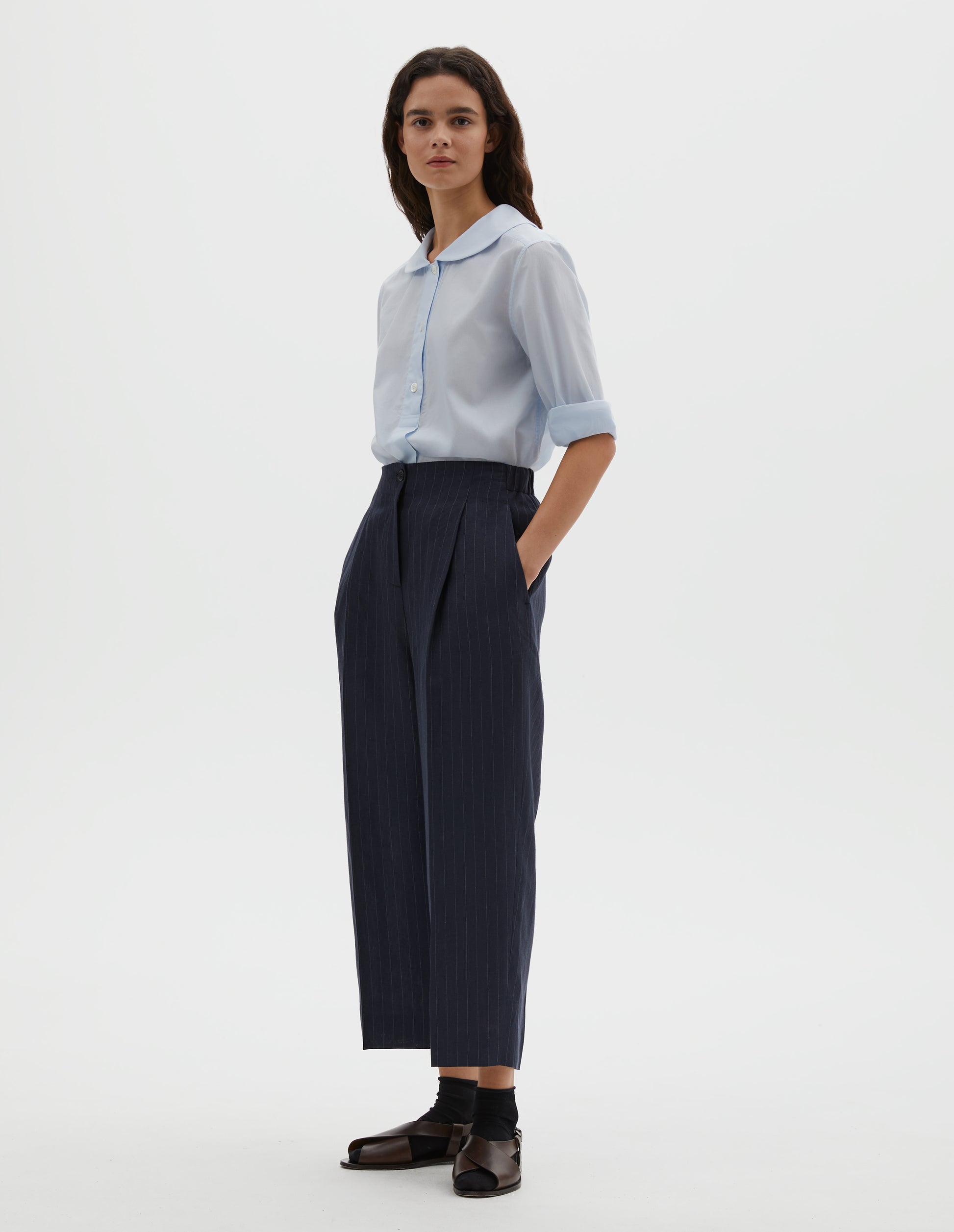 Full body image, front view of model wearing dark navy and grey pinstripe linen trousers, with horn button fastening, single pleats and slant side pockets. Styled with light blue petal collar shirt, black ankle socks and brown leather sandals.