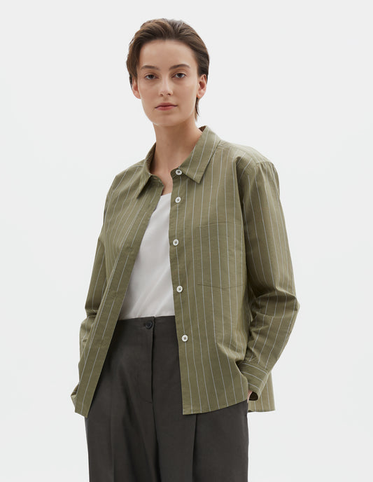 Cropped body image, front view of model wearing green with light blue pin stripe shirt with raised placket, mother of pearl buttons and patch pocket on chest. Styled over white t-shirt with brownish green trousers.