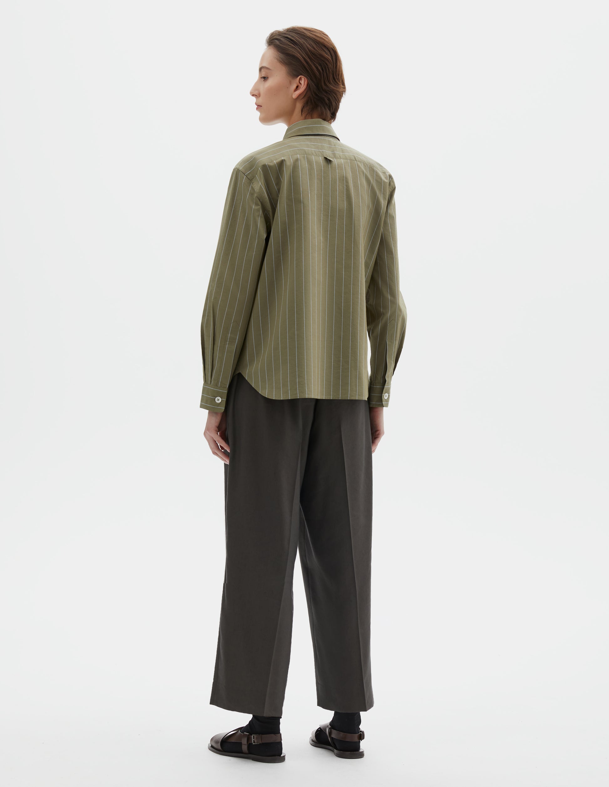 Full body image, back view of model wearing green with light blue pin stripe cotton silk shirt with mother of pearl buttons on cuffs and hanger loop detail on back yoke. Styled with brownish green trousers and black socks with dark brown leather sandals.