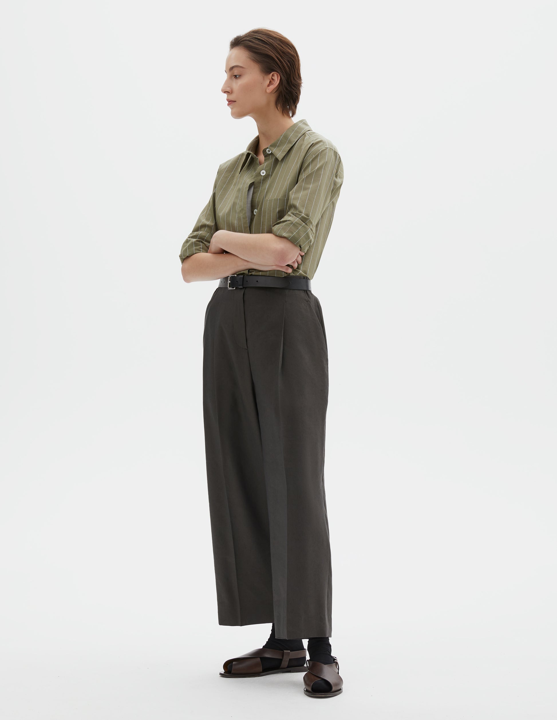 Full body image, front view of model wearing brownish green linen silk trousers, with horn button fastening, single pleats and slant side pockets. Styled with green and pale blue pin stripe shirt, black ankle socks and brown leather sandals.