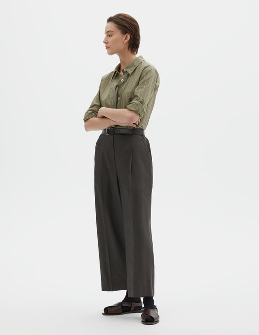 Full body image, front view of model wearing brownish green linen silk trousers, with horn button fastening, single pleats and slant side pockets. Styled with green and pale blue pin stripe shirt, black ankle socks and brown leather sandals.