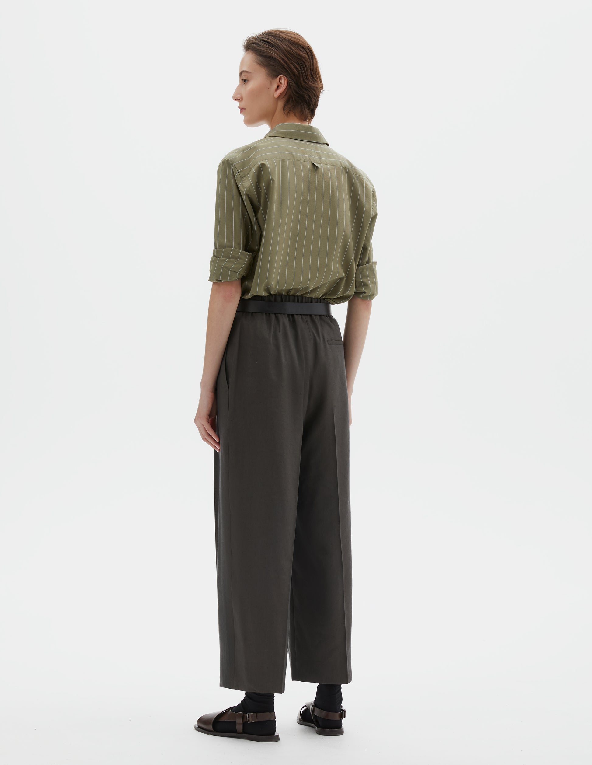 Full body image, back view of model wearing  brownish green linen silk trousers, with half elasticated waistband and belt back pocket. Styled with green and pale blue pin stripe shirt, black ankle socks and brown leather sandals.