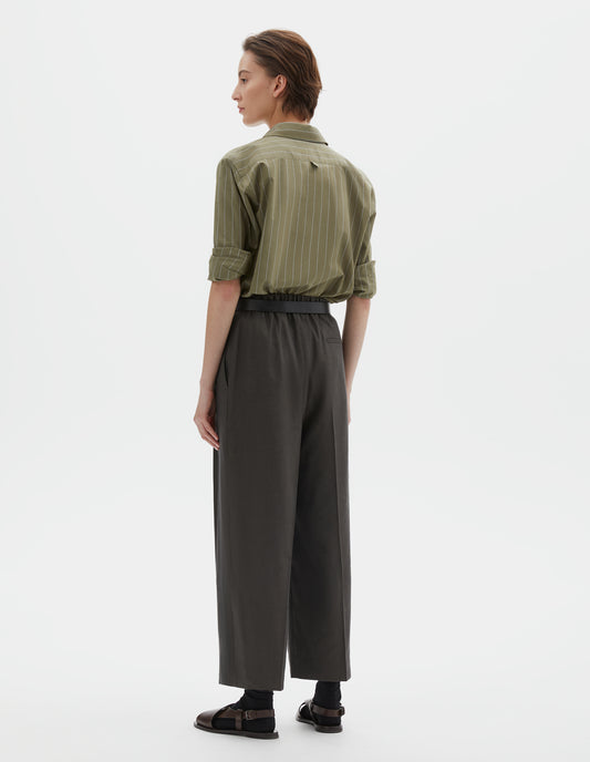 Full body image, back view of model wearing  brownish green linen silk trousers, with half elasticated waistband and belt back pocket. Styled with green and pale blue pin stripe shirt, black ankle socks and brown leather sandals.