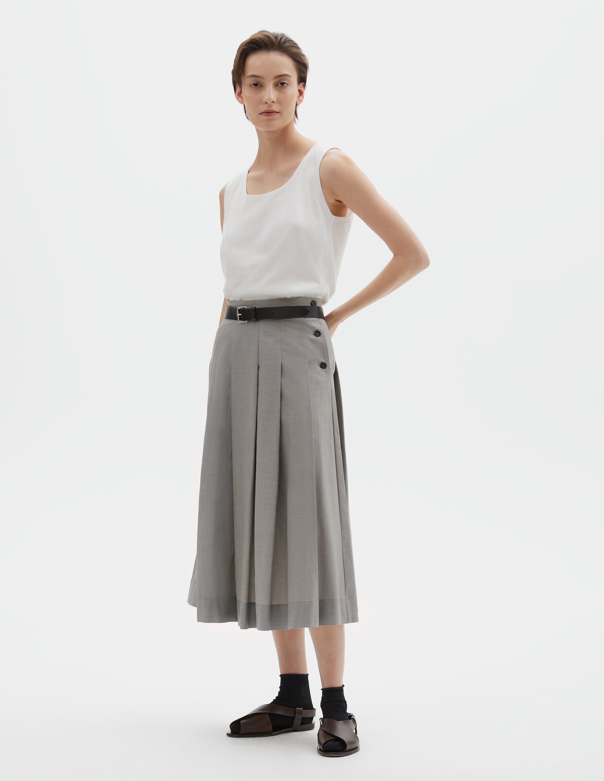 Full body image, front view of model wearing off white cotton sleeveless vest with scoop neck. Styled with grey pleated linen skirt and black leather belt, with black ankle socks and brown leather sandals.