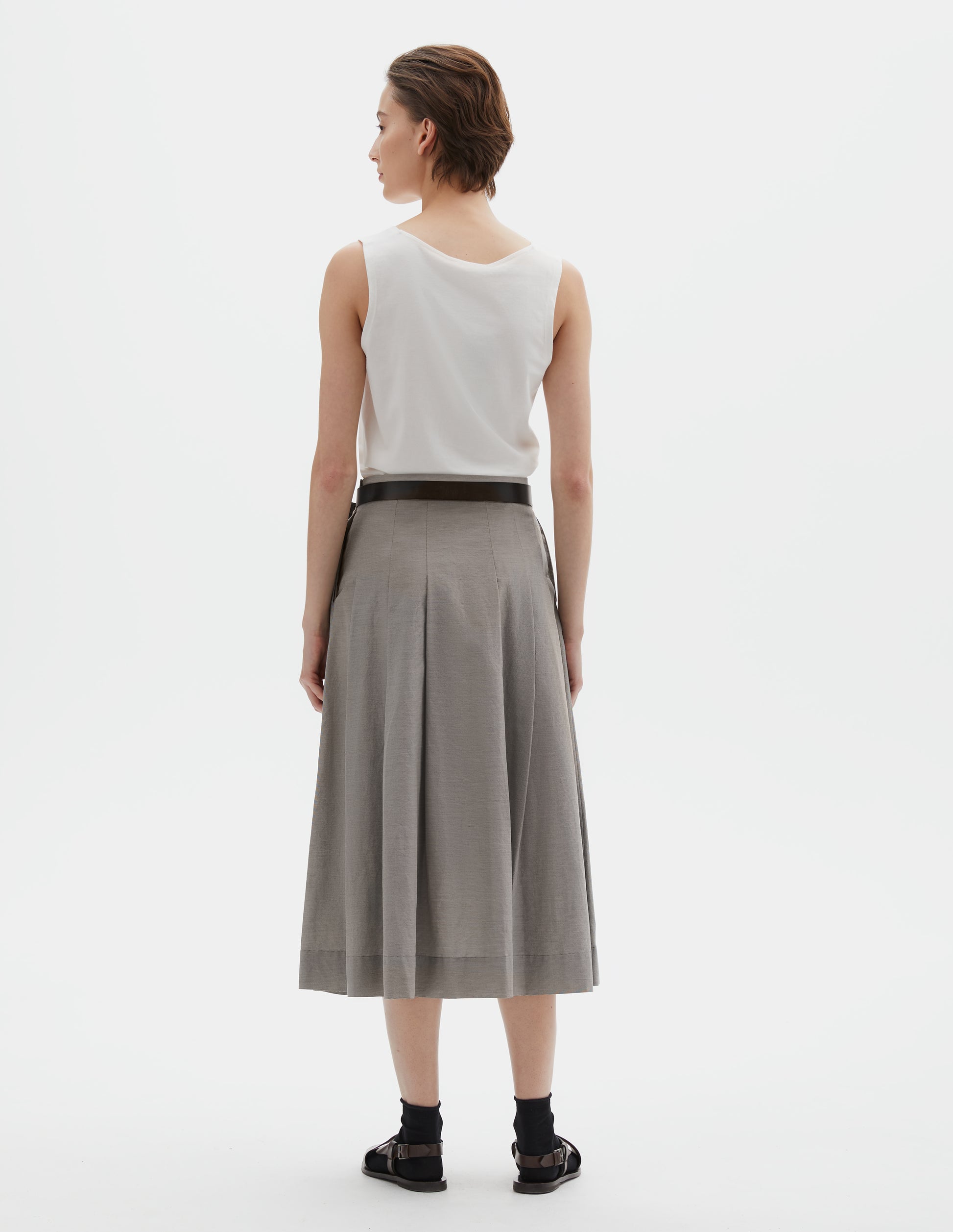 Full body image, back view of model wearing off white cotton sleeveless vest, Styled with grey pleated linen skirt and black leather belt, with black ankle socks and brown leather sandals.