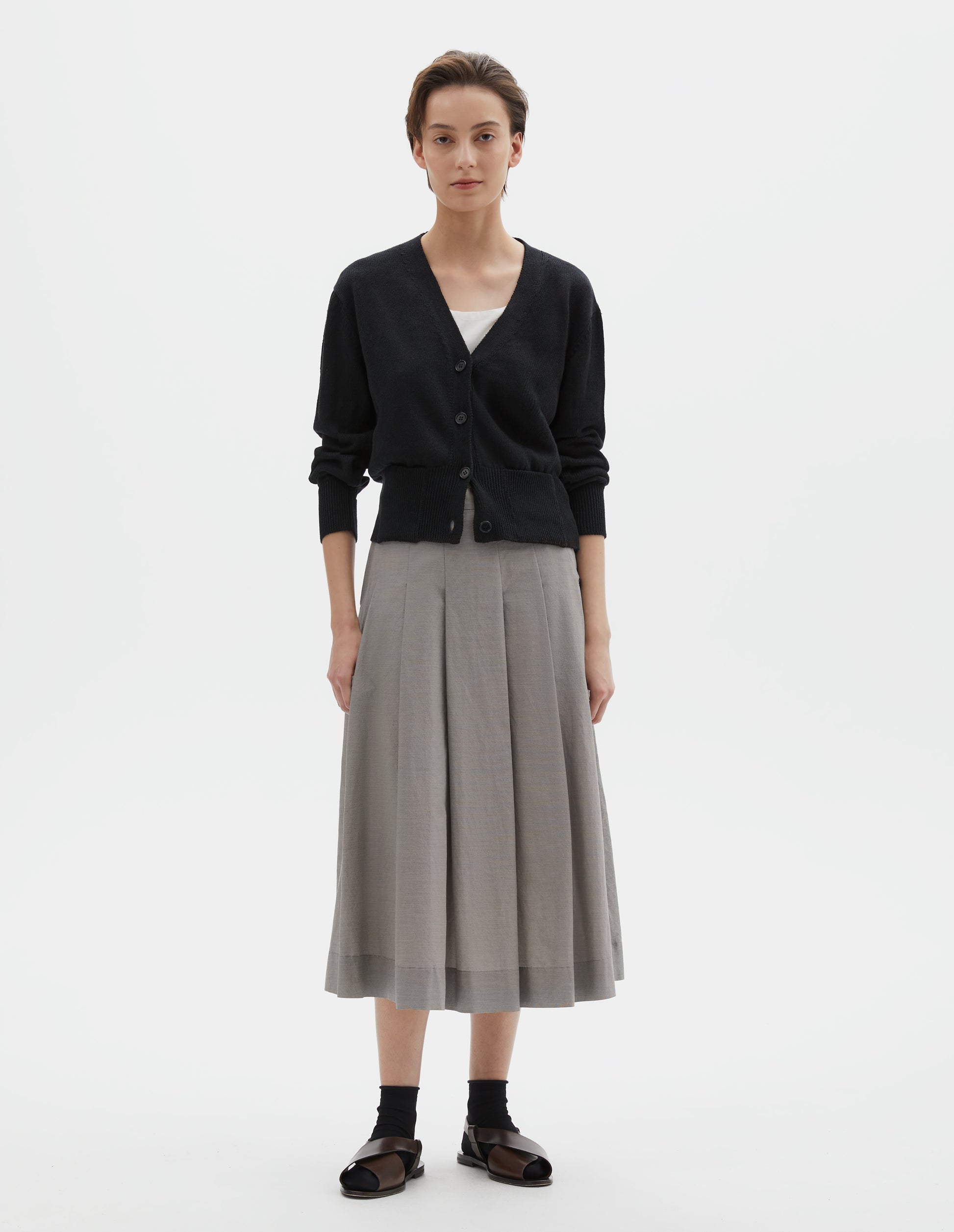 Full body image, front view of model wearing black linen cotton v neck cardigan with horn buttons, integral pockets and rib on neck, cuffs and wide hem. Styled with grey pleated skirt, black socks and black leather sandals.