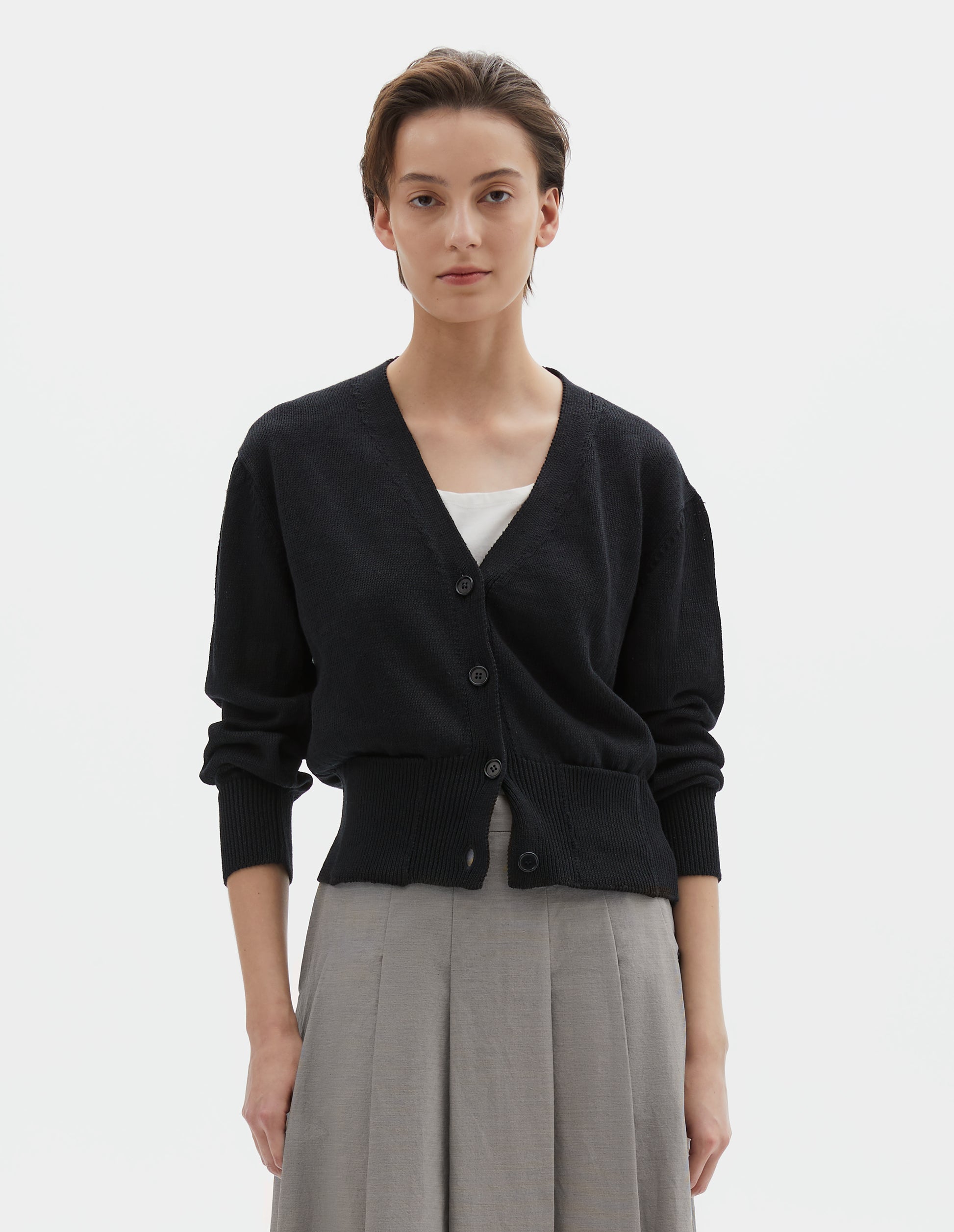 Cropped body image, front view of model wearing black linen cotton v neck cardigan with horn buttons, integral pockets and rib on neck, cuffs and wide hem. Styled with grey pleated skirt.