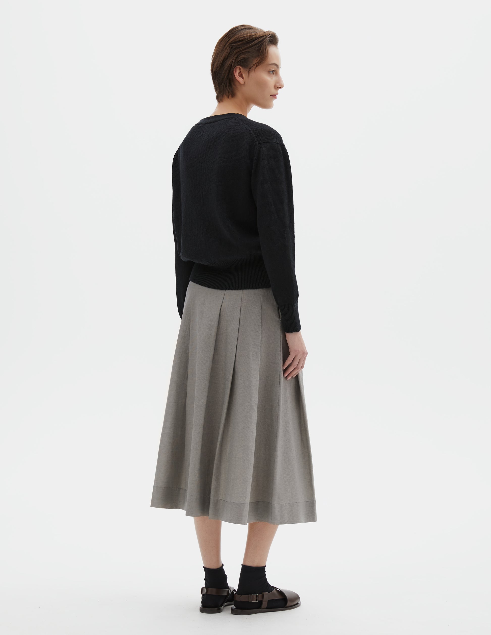 Full body image, back  view of model wearing black linen cotton cardigan with rib on neck, cuffs and wide hem. Styled with grey pleated skirt, black socks and black leather sandals.
