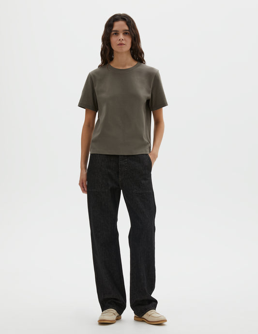 Full body image, front view of model wearing black denim twill jeans, with metal button fly, stitch-through waistband with belt loops, and stitch through pockets. Styled with green t-shirt and beige suede loafers. 