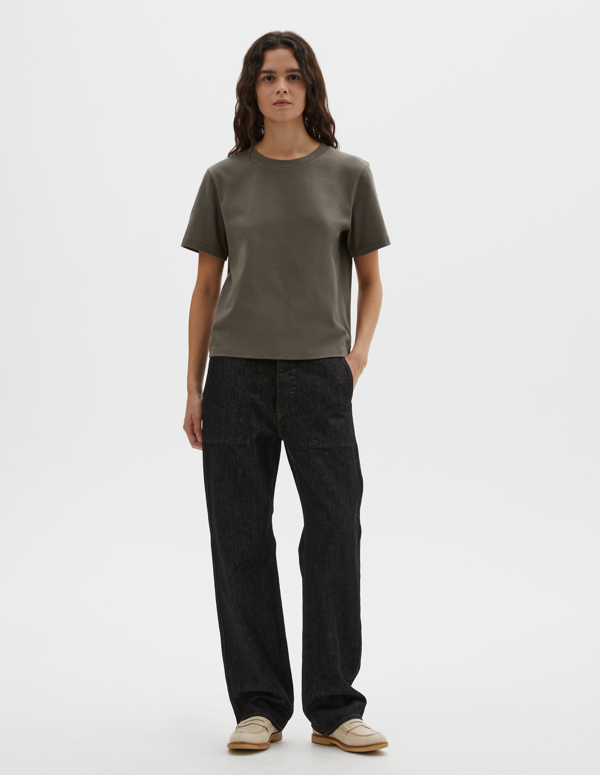 Full body image, front view of model wearing greyish green cotton short sleeve t-shirt with boxy cut. Styled with black denim jeans and beige suede loafers.