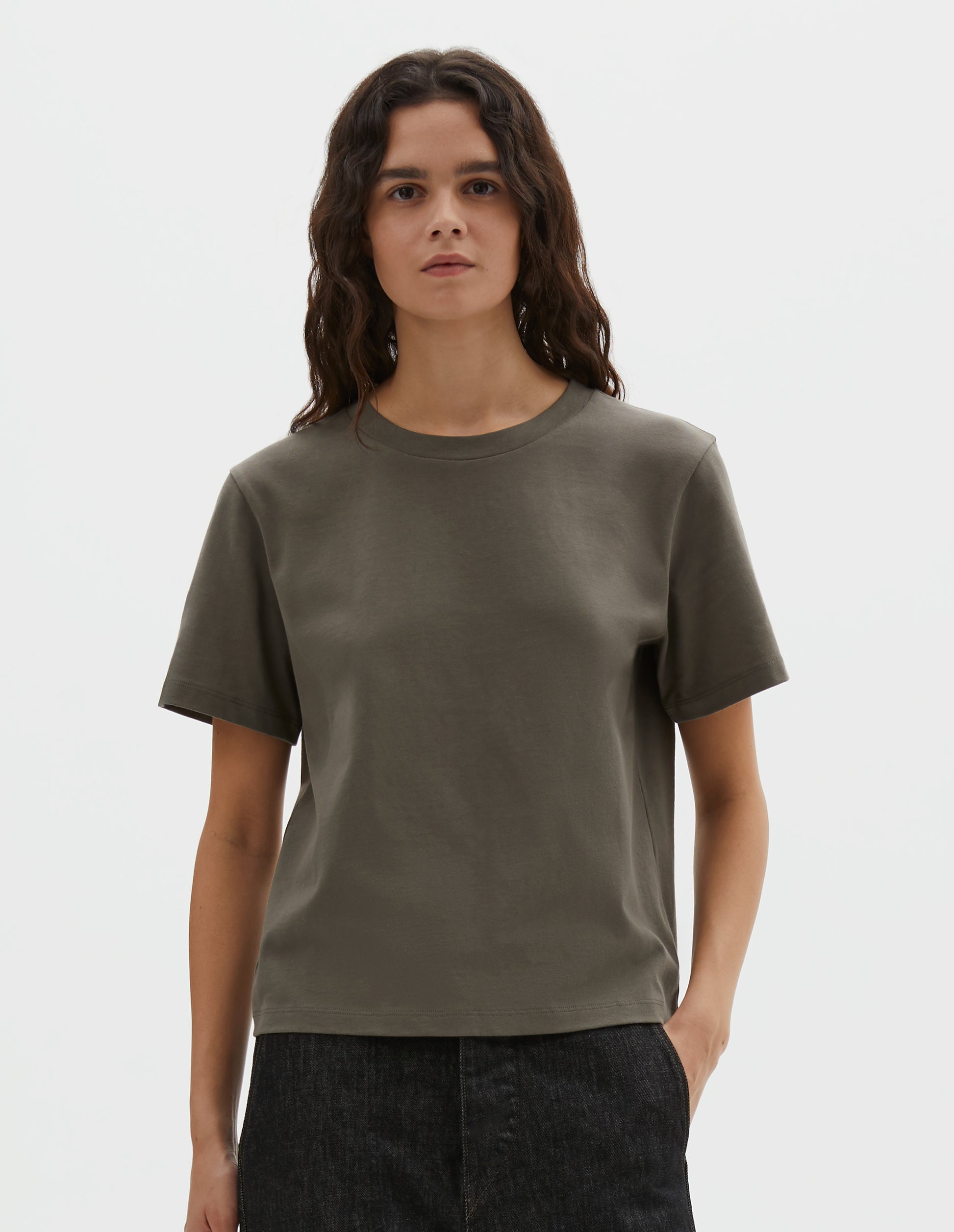 Cropped body image, front view of model wearing greyish green cotton short sleeve t-shirt with boxy cut. Styled with black denim jeans.