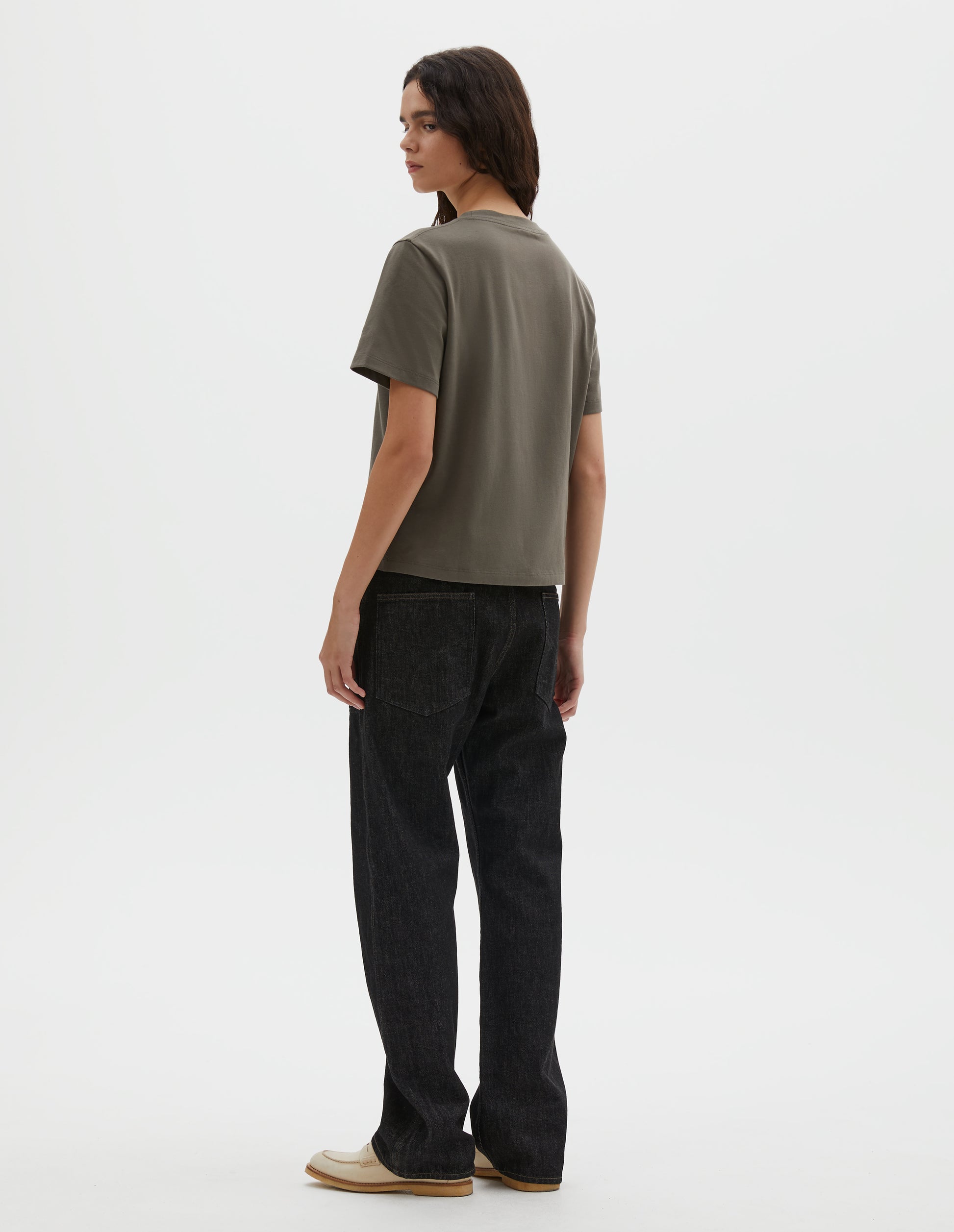Full body image, back view of model wearing black denim twill jeans, with stitch-through waistband with belt loops, cinch back detail with metal buckle and patch back pockets. Styled with green t-shirt and beige suede loafers. 