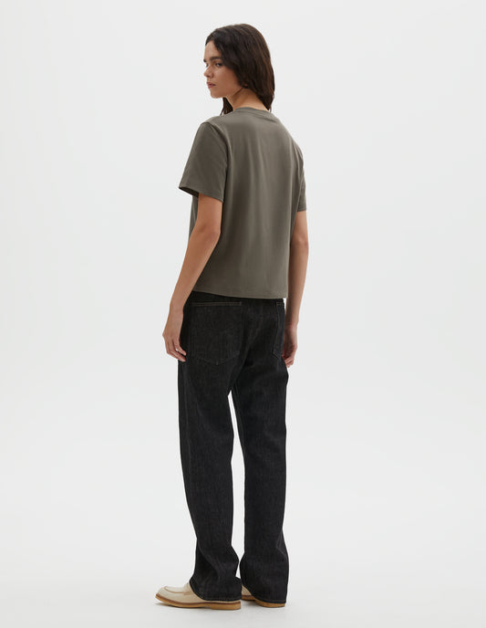 Full body image, back view of model wearing black denim twill jeans, with stitch-through waistband with belt loops, cinch back detail with metal buckle and patch back pockets. Styled with green t-shirt and beige suede loafers. 
