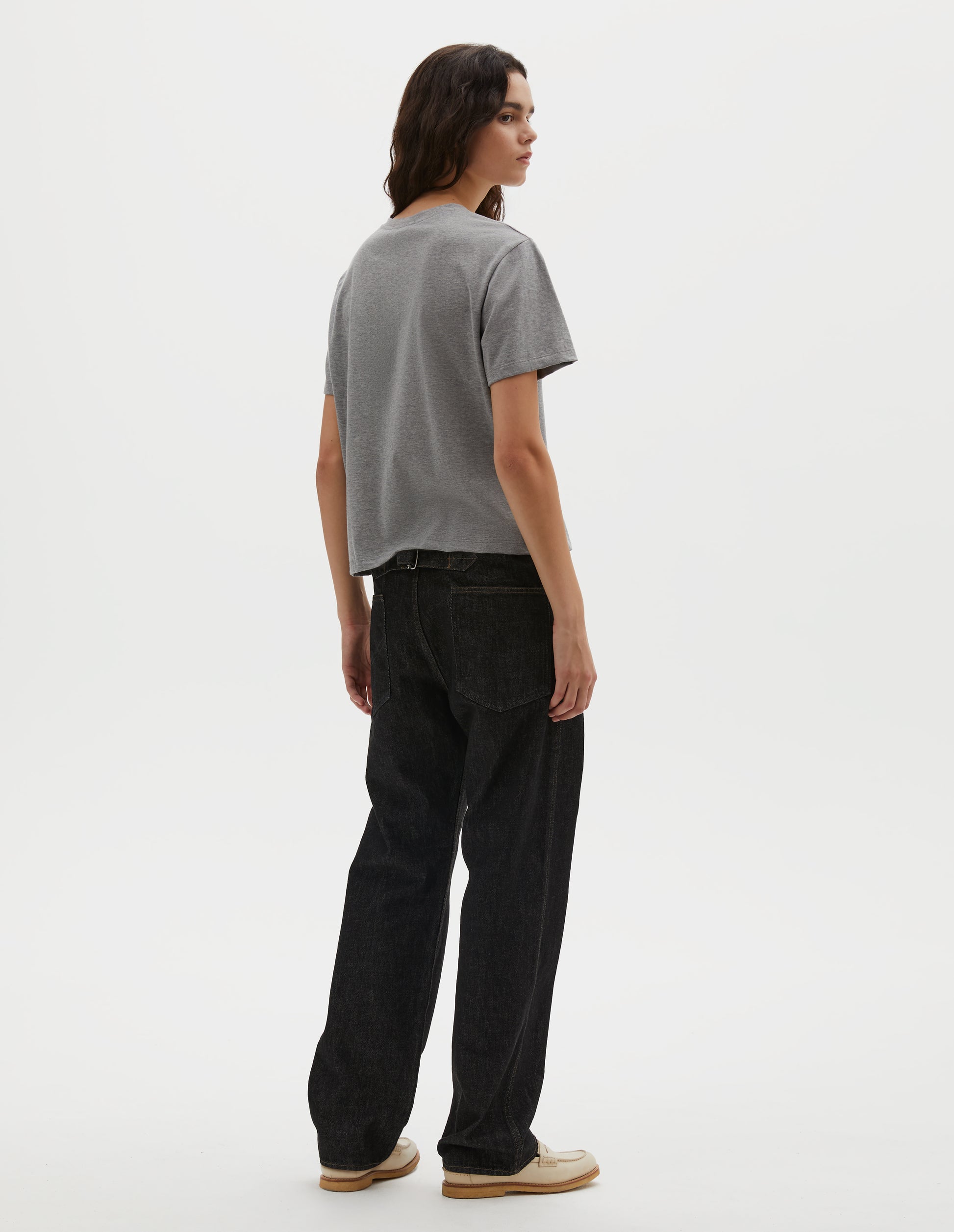 Full body image, back view of model wearing grey cotton short sleeve t-shirt with boxy cut. Styled with black denim jeans and beige suede loafers.