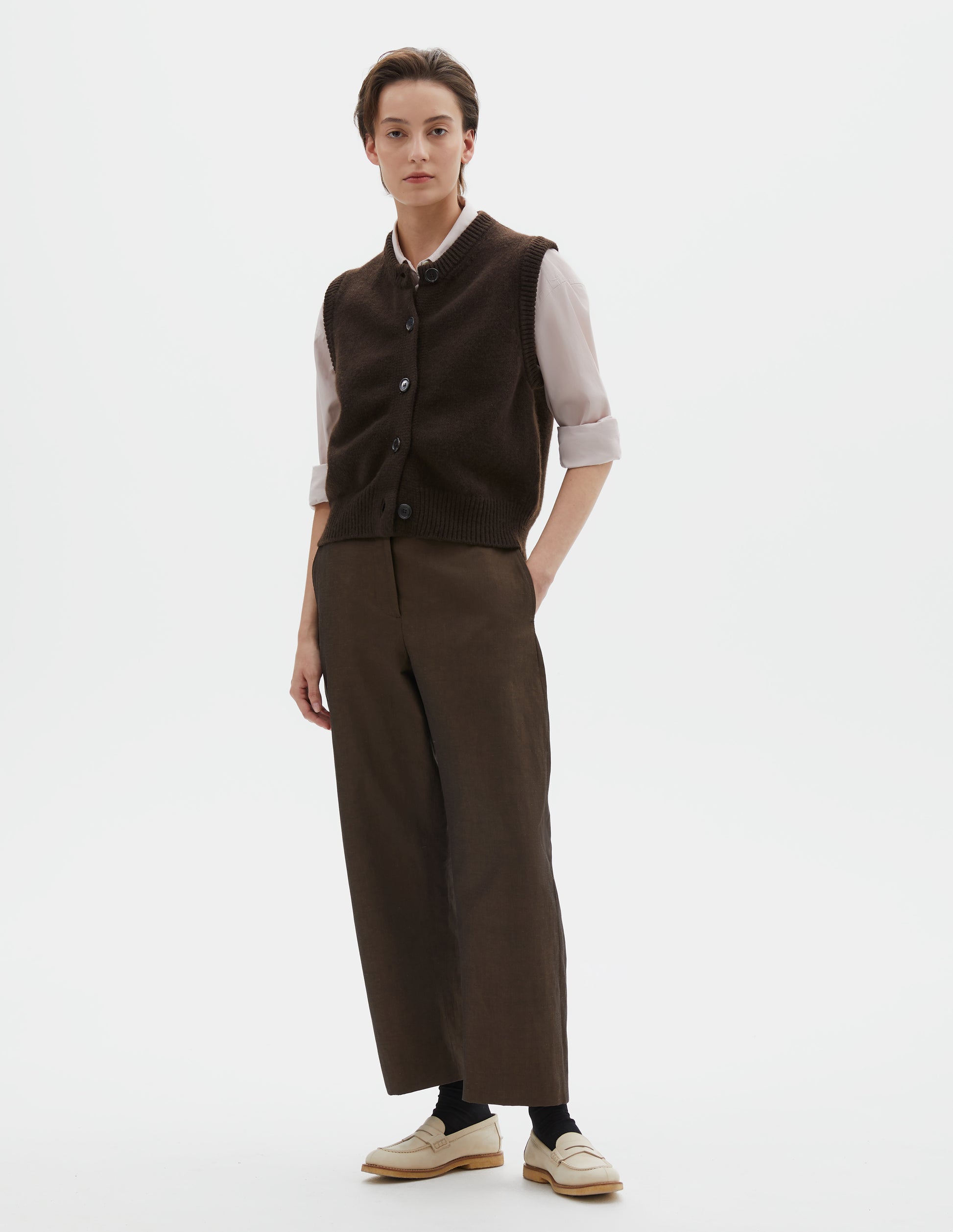 Full body image, front view of model wearing dark brown sleeveless merino cashmere gilet with contrast rib neckline, hem and dark horn buttons. Styled with pink shirt and brown trousers, black socks and beige shoes.