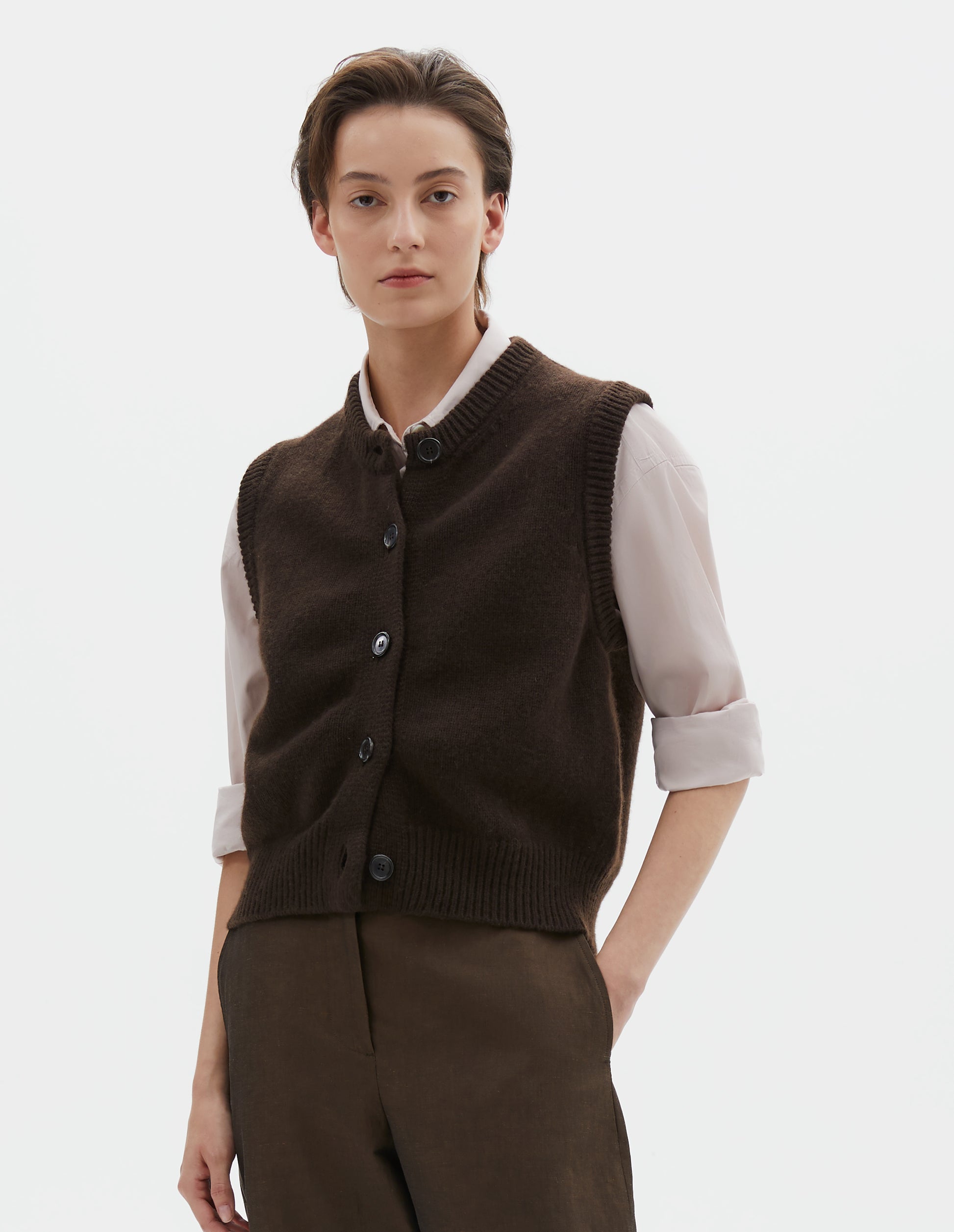 Cropped body image, front view of model wearing dark brown sleeveless merino cashmere gilet with contrast rib neckline, hem and dark horn buttons. Styled with pink shirt and brown trousers.