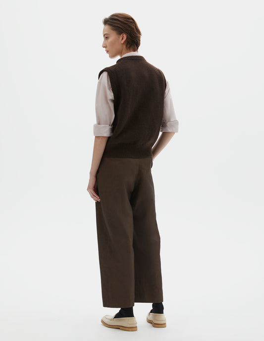 Full body image, back view of model wearing  dark brown sleeveless merino cashmere gilet with contrast rib neckline and hem. Styled with pink shirt and brown trousers, black socks and beige shoes.