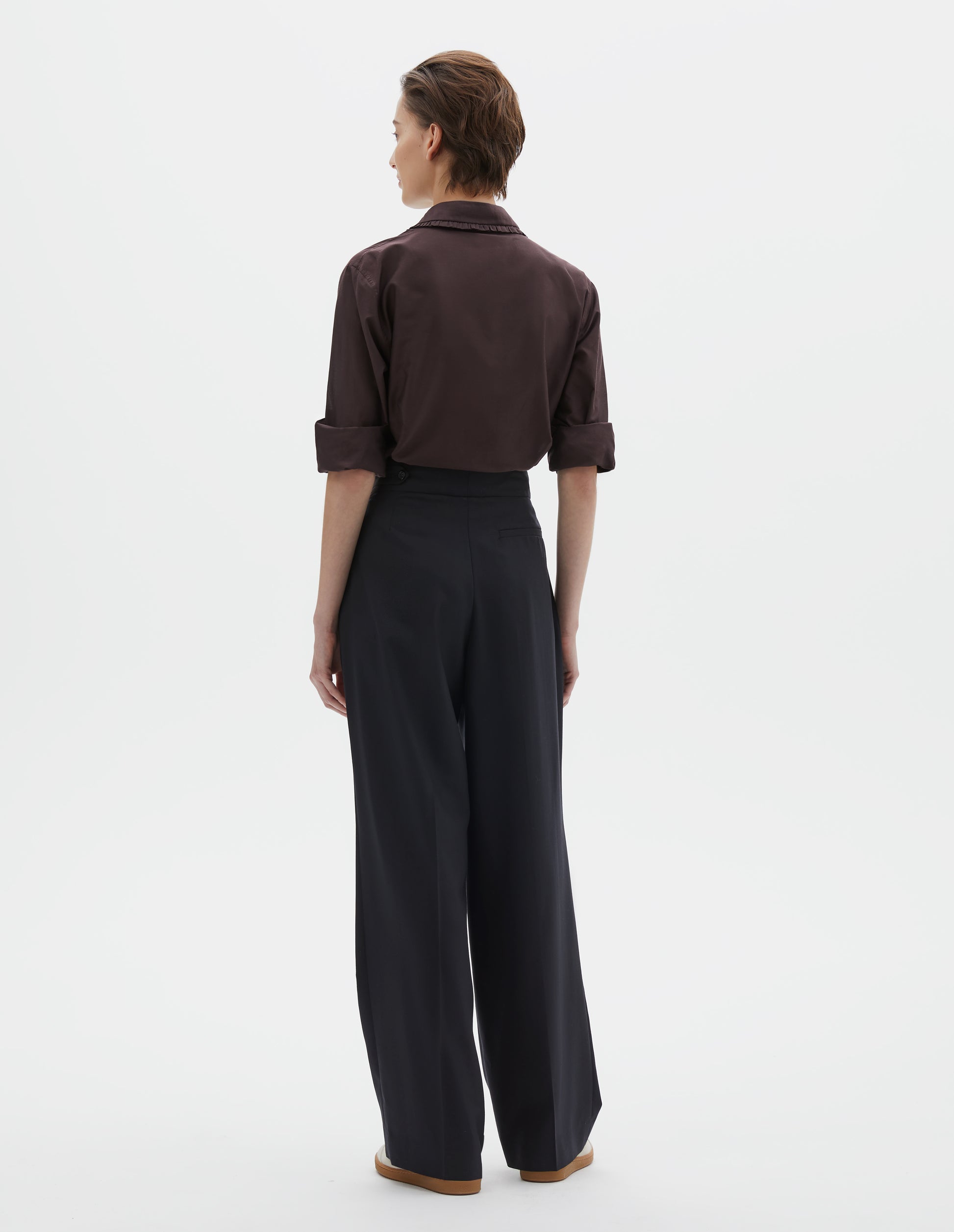 Full body image, back view of model wearing dark navy summer wool trousers with pull through double tab side fastening and back pocket. Styled with brown shirt and beige trainers.
