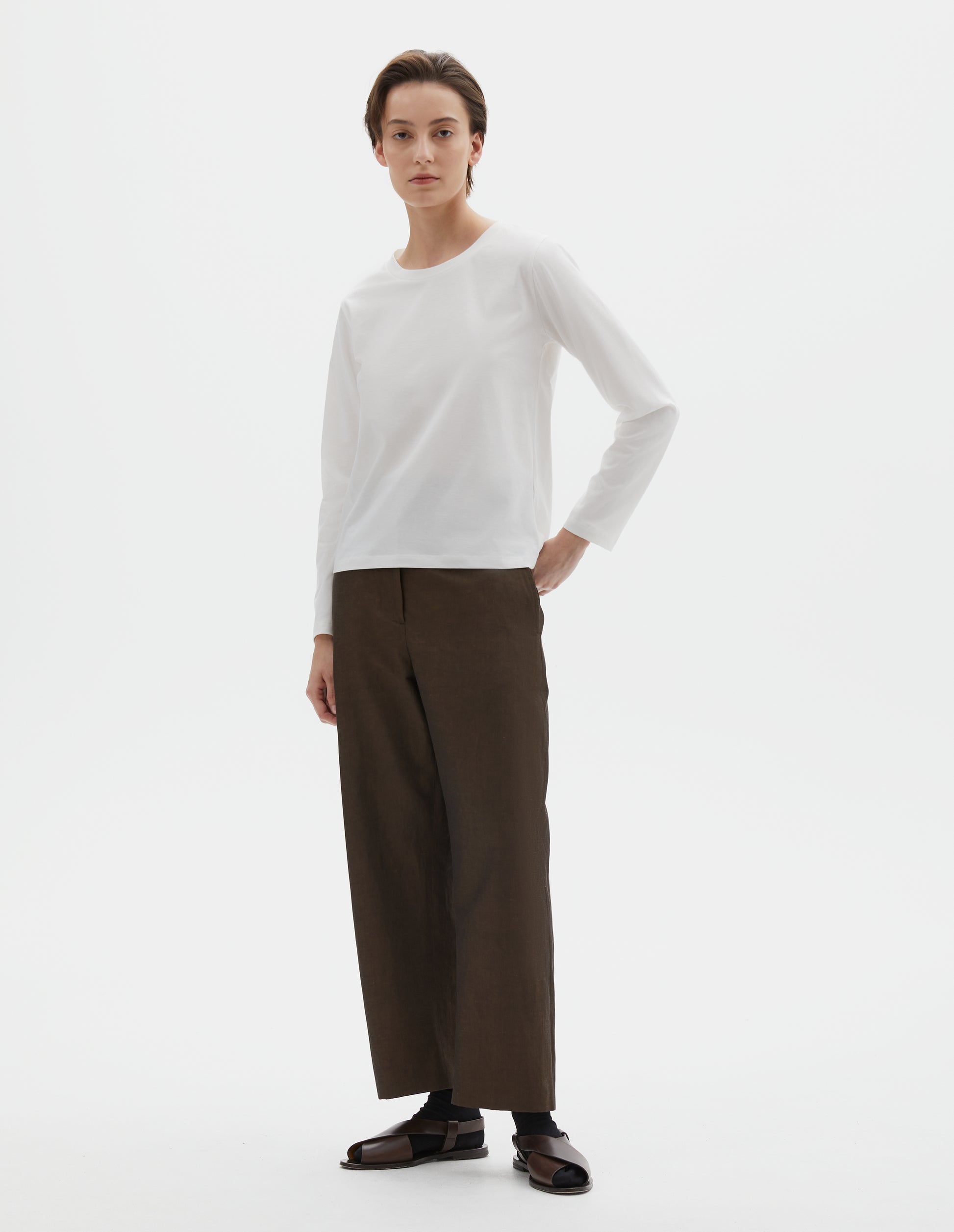 Full body image, front view of model wearing off white cotton wide neck long sleeve with boxy cut. Styled with brown trousers, black socks and brown leather sandals.