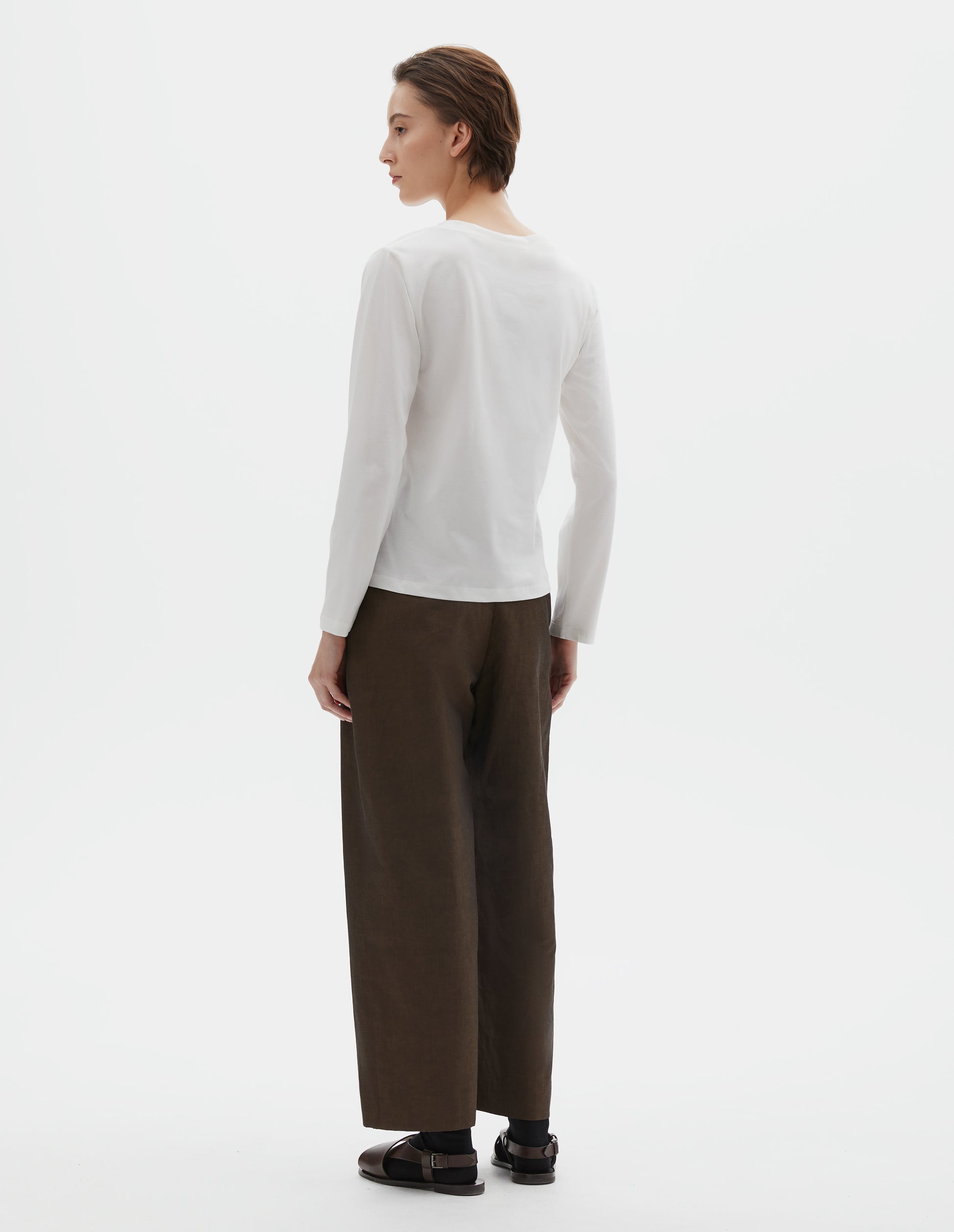 Full body image, back view of model wearing off white cotton wide neck long sleeve with boxy cut. Styled with brown trousers, black socks and brown leather sandals.