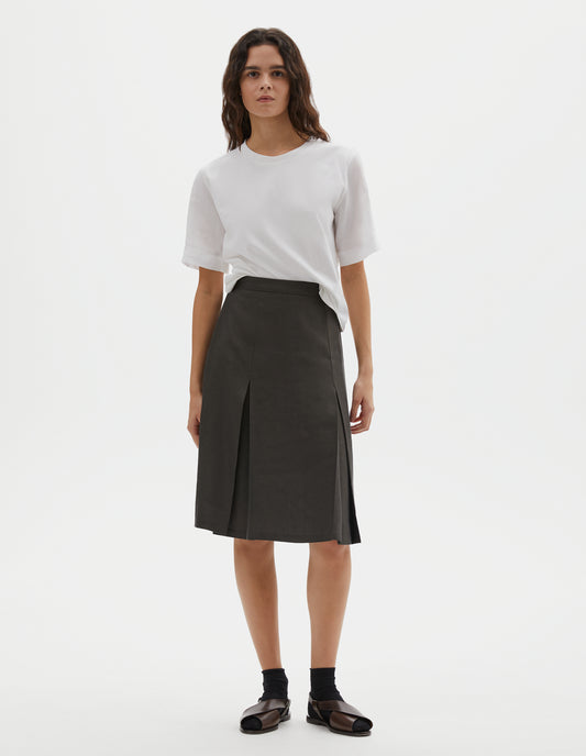 Full body image, front view of model wearing greenish brown regular A line knee length skirt, minimal waistband with concealed side zip and full hem. Styled with white t-shirt, black socks and brown leather sandals.