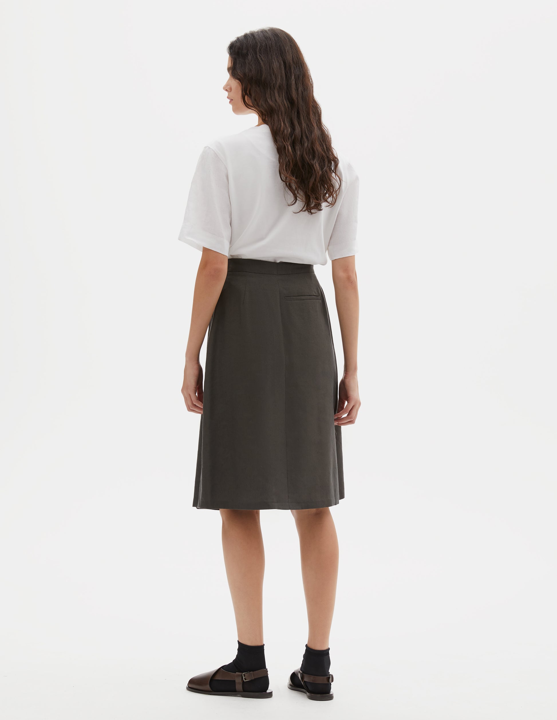 Full body image, back view of model wearing greenish brown regular A line knee length skirt, minimal waistband with jet pocket and full hem. Styled with white t-shirt, black socks and brown leather sandals.