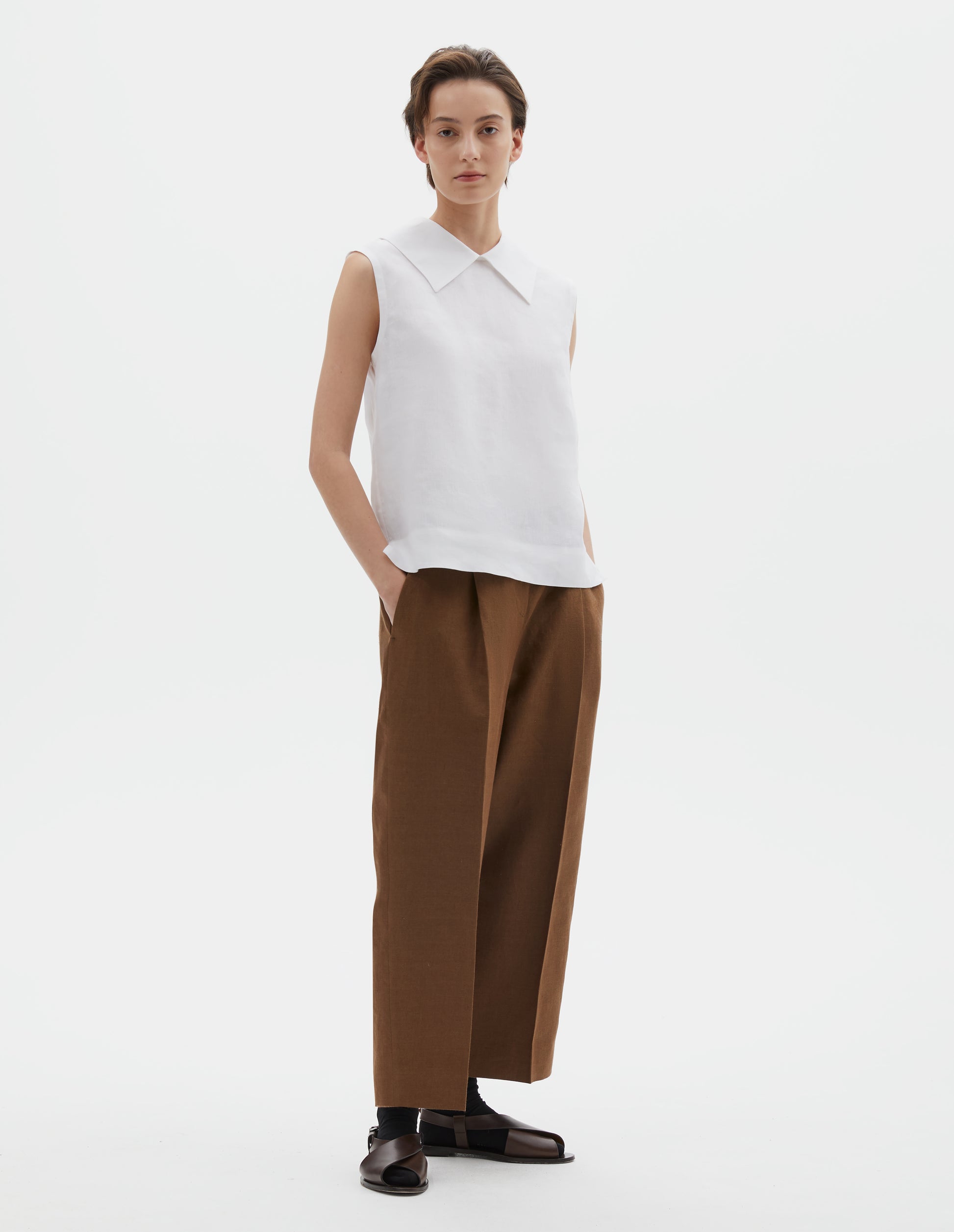 Full body image, front view of model wearing white regular cut Irish linen top with exaggerated collar, side dart detailing, with a deep hem with side splits. Styled with brown trousers, black socks and brown sandals.
