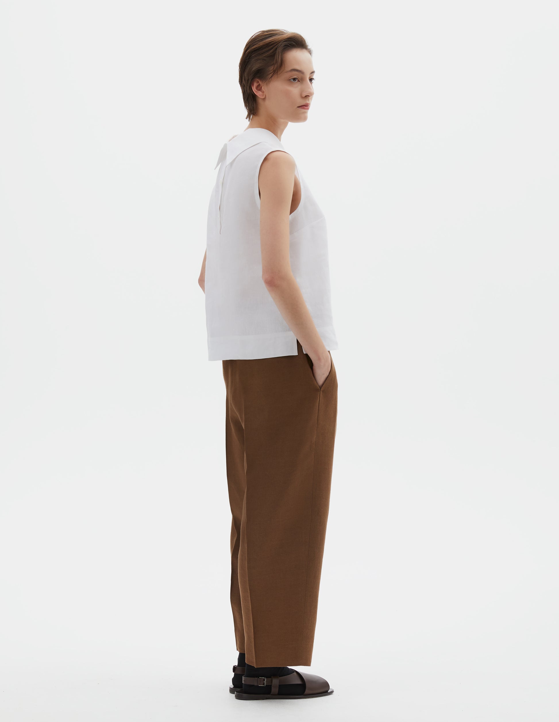 Full body image, back view of model wearing white regular cut Irish linen top with exaggerated collar, button-fastening placket at the centre back, with a deep hem with side splits. Styled with brown trousers, black socks and brown sandals.